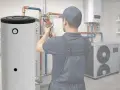 Instalator pracuje przy dużym, pionowym wymienniku CWU 200l. W tle widać pompę ciepła i inne elementy instalacji.