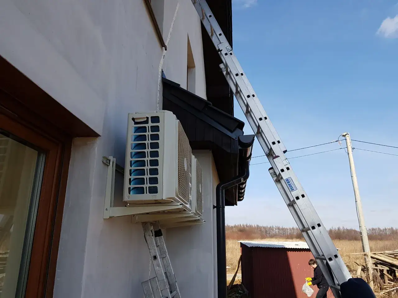 Montaż jednostki zewnętrznej klimatyzacji Rotenso Split na ścianie budynku. Drabina oparta o dom, w tle niebo i pola.