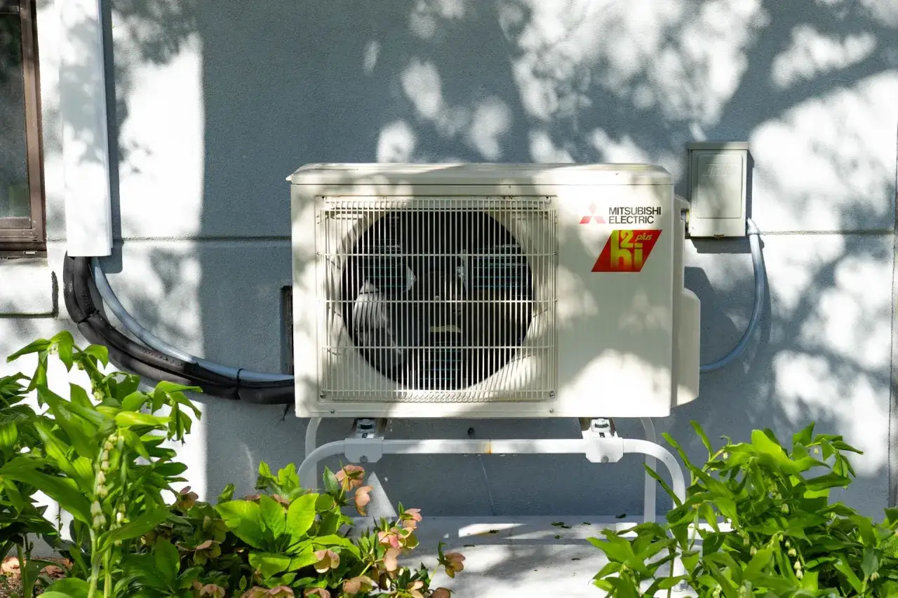 Pompa ciepła bez fotowoltaiki: czy się opłaca? Jednostka zewnętrzna klimatyzatora Mitsubishi Electric na ścianie budynku, otoczona zielenią.